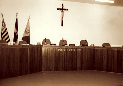 Plenário, bancada de madeira com cadeiras. No canto esquerdo bandeiras do estado, município e país. Crucifixo ao centro da parede ao fundo.